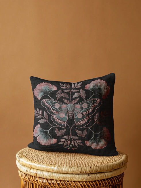 A square decorative pillow featuring an intricately detailed Luna moth in faded rose pink, slate blue and dusty terracotta colors on a black background. The moth is centered in a symmetrical arrangement with ginkgo-inspired leaves and botanical elements surrounding it.